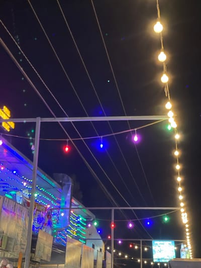 This night-time image captures a vibrant outdoor scene in Kimorori, Kenya, likely depicting a market, fair, or public event space. The dark sky is dramatically lit by an array of artificial lights.
A prominent string of warm yellow/white globe lights stretches horizontally across the upper portion of the frame, supported by wires and poles. Below this, numerous colorful LED lights, in shades of blue, purple, red, and green, are strung vertically and horizontally, creating an intricate web of illumination. These lights are attached to multiple white poles and thin wires that crisscross the frame.
On the right side and in the lower half of the image, structures resembling market stalls or temporary buildings are visible, also adorned with colorful lights. These structures have angular designs, with some surfaces appearing to be made of corrugated material or draped fabrics. In the upper right quadrant, a brightly lit rectangular screen or display is visible, showing indistinct content. The extensive lighting and temporary structures suggest a lively commercial or festive gathering.
The scene is clearly set at night, with the artificial lights being the primary source of illumination against the dark background. No individuals are visible, but the setup implies a bustling public event. The overall impression is one of festivity and activity. There is no discernible text visible in the image.