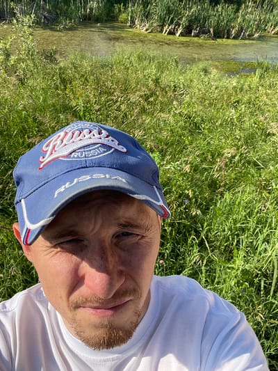 This is a close-up, horizontal, selfie-style photograph of a man wearing a blue baseball cap and a white t-shirt. The man's face is visible, with his eyes closed and his head tilted. He has short, light brown hair and stubble on his face. The cap has an embroidered logo with white and red detailing, and some text is visible on the brim.
The man is outdoors, sitting or lying on grass. Behind him, to his left, is a body of water, possibly a stream or river, bordered by lush green reeds and grass. The water appears shallow and clear. The overall setting suggests a natural, possibly rural environment. The sunlight is bright, casting shadows and indicating it is daytime, likely late morning or afternoon. The weather appears to be warm and sunny.
Visible text on the cap includes "RUSSIA" and what appears to be "FEDERATION". The scene evokes a sense of relaxation and enjoyment of the outdoors. The image is oriented sideways, requiring rotation for proper viewing.