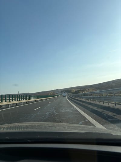 The image presents a forward-facing perspective from inside a vehicle traveling on a multi-lane asphalt highway, identified as likely the A2 highway in the vicinity of Cernavoda, Romania. The vehicle's dashboard is visible across the bottom of the frame. The road surface features distinct white lane markings and irregular, scattered white streaking. Metal guardrails flank both sides of the highway; the guardrail on the left incorporates green vertical support posts. The surrounding landscape consists of gently sloping terrain or hillsides with sparse vegetation, including visible rows indicative of cultivation. Two vehicles are observed ahead in the distance: a white sedan in the rightmost lane, and a larger white vehicle further ahead, approaching what appears to be an overpass or tunnel entrance. The sky is clear and light blue, with a faint contrail visible in the upper right. The scene is illuminated by daylight, and shadows cast by the right guardrail suggest the sun is positioned to the left of the direction of travel. No individuals are explicitly visible.