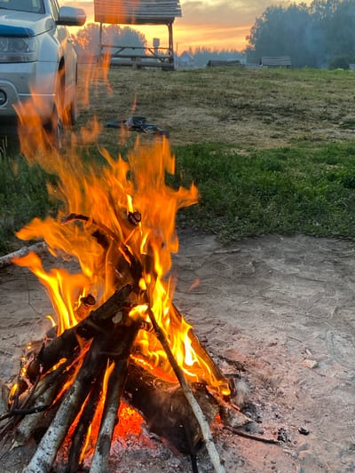 The image depicts a campsite at dusk in the vicinity of Peshcherka, Russia. A campfire burns brightly in the foreground, with logs smoldering and flames leaping upwards. To the left, the side of a silver SUV is visible. In the background, a wooden canopy structure stands on a grassy field. Beyond that, a line of trees marks the horizon, silhouetted against a soft, orange-hued sky. The ground is a mix of dirt and short, dry grass. There are no people visible in the frame.