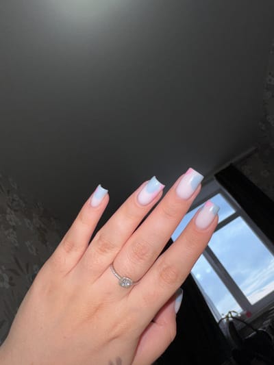 The image presents a close-up view of a person's right hand, likely taken indoors in Shirkovo, Russia. The hand features a detailed manicure with square-shaped nails adorned with a modern French tip design. The nail tips display a soft, blended gradient of pastel pink and light blue over a sheer, milky white base. A silver-toned ring, featuring a central clear gemstone (possibly square or cushion cut) surrounded by smaller pavé-set stones on the band, is prominently worn on the ring finger.

The hand is positioned against a background that includes a window revealing an overcast or cloudy sky, indicating that the photograph was taken during daytime with natural, diffused light. To the left, a dark, plain wall or surface is visible, while the bottom right corner shows a dark surface with a faint, subtle floral or abstract pattern, possibly a piece of furniture or fabric. The overall composition suggests the hand, manicure, and ring are the primary subjects being showcased. There is no visible text in the image.
