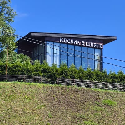 The image shows a modern building with large glass windows and a dark brown exterior, situated on a grassy hillside. White text reading "КРОЛИК В ШЛЯПЕ" is visible on the facade of the building. The building is surrounded by lush green bushes and trees. A rustic wooden fence runs along the base of the hill. Overhead power lines are visible in the foreground. The sky is clear and blue. The scene appears to be in a natural, semi-rural setting, consistent with the location of Yanino-2, Russia. There are no people visible in the image.