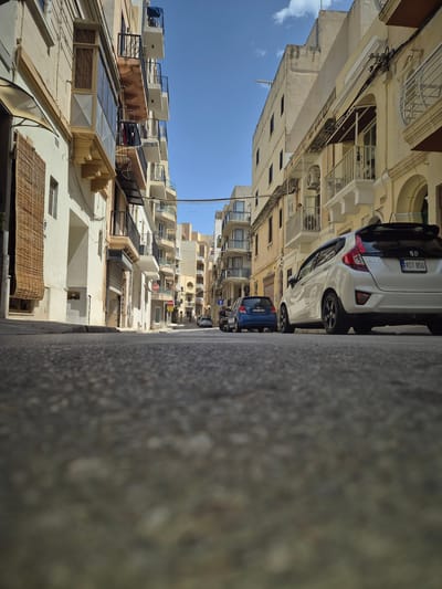 This is a low-angle, eye-level shot of a narrow street in Pietà, Malta, on a sunny day. The street is paved with asphalt and shows some wear and tear. The buildings lining the street are typical of Maltese architecture, with light-colored facades, balconies with wrought-iron railings, and many windows. The buildings are several stories tall and appear to be residential or commercial.
In the foreground, the asphalt is detailed, showing texture and small stones. Two cars are parked on the right side of the street. The closest car is a white Honda Fit, facing away from the camera, with a license plate that reads "RIT 850". Behind it, a blue hatchback is also parked, facing away, with a license plate that appears to read "KJT 003". A dark motorcycle is parked to the left of the blue car. Further down the street, more buildings and parked cars are visible, creating a sense of depth and perspective. The sky is a clear, bright blue with a few white clouds. The sunlight casts shadows on the street and buildings, indicating a bright, sunny day. A few electrical wires are visible strung between the buildings. A "STOP" sign is visible on a pole on the right side of the street in the mid-ground. The overall impression is of a typical urban scene in Malta, with a focus on the architectural details and the presence of parked vehicles.