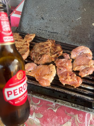 In Stornara, Italy, a grill is laden with an assortment of meats, including several pieces of raw chicken and two cuts of steak. The chicken pieces, varying in size and shape, are arranged on the lower grates of the grill, appearing marinated with spices. Above them, two seared steak portions are positioned on the upper grates, showing distinct grill marks. In the foreground, a partially visible bottle of Peroni beer, identifiable by its red and white label, is held by a hand, obscuring a portion of the grill and its contents. The background reveals a blurred pink and white box, and the grill is situated on a surface covered with a red tablecloth featuring a gold floral pattern. The lighting suggests an outdoor setting, possibly during daylight.