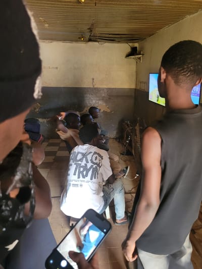 In Bokkos, Nigeria, a group of young men are gathered in what appears to be a common room or lounge area, likely a home or community space. They are engrossed in watching television, with several individuals seated and lying on the floor facing a TV screen mounted on the wall. The room has a rustic and somewhat unkempt feel, with exposed wiring, corrugated metal sheeting on one wall, and a visible fan. The lighting suggests it is daytime, though the interior is somewhat dim.
One man in the foreground is in profile, wearing a dark t-shirt, and his head is tilted upwards as if looking at the TV or something beyond the frame. Behind him, several other men are positioned on the floor, some lying down, others sitting up. One man is wearing a white t-shirt with a graphic design. Another is seen wearing a light-colored t-shirt. The overall atmosphere seems relaxed and communal, typical of friends or family spending time together.
In the lower right corner, a hand holding a smartphone is visible, capturing the scene. The phone screen displays a partial image of someone's legs and feet. The text "BOSS-MAN" and "ESTI" are visible on the wall in the background. The text "طيران الرياض" (which translates to "Riyadh Air") is seen on the television screen, suggesting a sports broadcast or news program. The image conveys a sense of casual leisure and shared experience.
