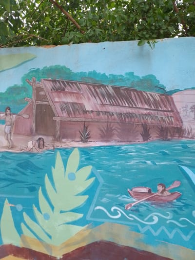 The image is a section of a mural, possibly located in the city of Villa Hayes, Paraguay. The mural depicts a scene of indigenous life, possibly representing the pre-colonial era. The primary subject is a large thatched structure, resembling a traditional dwelling, situated on a sandy shore adjacent to a body of water. Three human figures are visible: one is wading or swimming in the water, another is on land carrying what appears to be a tool or weapon over their shoulder, and a third is paddling a small boat. The water is a stylized depiction of waves and floral designs, with geometric patterns also present. Above the mural, foliage and tree branches are visible, indicating an outdoor setting. The mural has an overall painted style with some signs of wear and age.