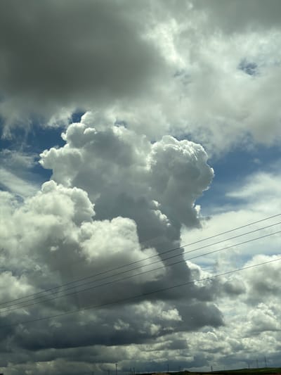 The image depicts a sky over Kiryat Gat, Israel, characterized by a dynamic cloud formation. A large, bright white cumulus cloud dominates the central and right portions of the frame, exhibiting significant vertical development and complex, textured surfaces illuminated by sunlight. Surrounding and underlying this formation are darker, greyish cloud masses, particularly in the upper left and lower sections, suggesting varying cloud thickness and light conditions. Patches of clear blue sky are visible between the clouds, predominantly on the right side.

In the mid-ground, four dark-colored power lines stretch horizontally across the frame, slightly descending from left to right. These lines exhibit minor reflections, suggesting the image was captured through a glass pane. At the distant horizon, a subtle landscape of undulating terrain is visible, with hints of green vegetation and reddish-brown earth. Several utility poles or electrical transmission towers are faintly discernable along this distant horizon line. No individuals, animals, or explicit actions are present within the scene.