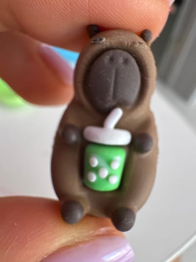 A macro photograph shows a small, brown, inanimate figurine of a capybara wearing a hoodie and holding a green cup with a white straw. The figurine has a dark brown nose and two small, dark brown ears. The cup has white dots on its exterior. The figurine is held between two human fingers, one on the top with visible fingerprints and the other on the bottom. The background is blurred and appears to be a white surface with hints of blue and green. The image is an extreme close-up, with the figurine taking up most of the frame. No location in Uyutnoye, Ukraine, is visible.