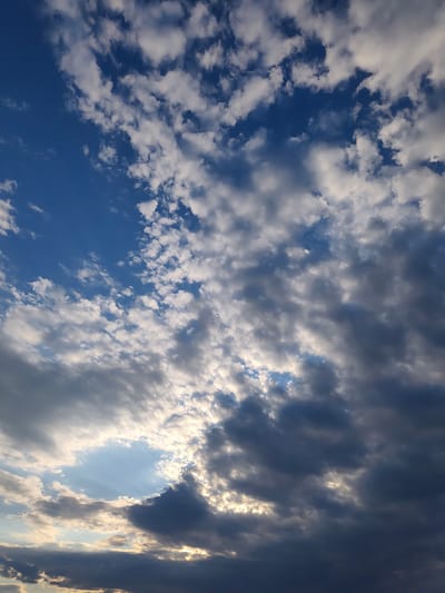 This is a photograph of a sky filled with clouds, viewed from the city of Codru, Moldova. The sky is predominantly blue with scattered white and dark gray clouds. Some of the clouds are illuminated by sunlight, creating bright patches and highlighting the textures of the cloud formations. The image appears to be taken during the daytime, possibly late afternoon or early evening, given the lighting. There are no people, buildings, or other man-made objects visible in the frame. The focus is entirely on the natural elements of the sky.