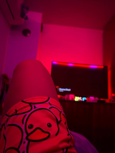 The scene depicts an indoor environment in Debelt, Bulgaria, illuminated predominantly by strong red and pink ambient lighting. In the foreground, the bare upper thigh of an individual is centrally positioned, while the lower torso/upper thigh area is covered by patterned clothing featuring repeating yellow cartoon ducks with black outlines and orange beaks. To the upper left, a white wall-mounted air conditioning unit is visible, with a small, dark blue, indistinct object attached to the wall or ceiling above it. Below the air conditioner, a circular dark object is affixed to the wall. In the mid-ground, a dark, rectangular flat-screen television is centrally located, with a strip of multi-colored LED lights (red and purple) illuminating the wall directly behind its upper edge. Below the television, on a dark surface, several indistinct objects emit glows of green, blue, and pink light. The surrounding walls and ceiling appear uniformly colored in shades of red and pink due to the ambient lighting. No individuals are fully visible.