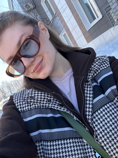 This close-up, daylight outdoor image depicts an individual, likely female, captured in a selfie in Новое Шигалеево, Russia. The subject wears large brown-framed sunglasses, a light pink inner shirt, a dark brown zippered jacket, and an outer vest or jacket featuring a black and white houndstooth pattern with horizontal black and white striped bands. A green strap, likely from a bag, crosses diagonally over the subject's chest. The subject has long, light brown hair, visible behind the left ear, and exhibits a subtle smile. The background reveals a modern multi-story building with a facade clad in alternating light brown/beige and dark grey/silver panels, featuring white-framed windows and an external air conditioning unit. Bare tree branches are visible to the left, and patches of white snow are present on the ground, indicating a cold weather season. The sky is clear.