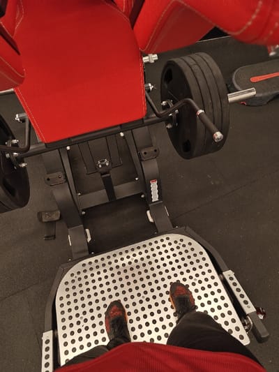 The scene depicts a gym environment in Nahariya, Israel, from a first-person perspective looking down at a leg press or hack squat machine. The lower torso, legs, and feet of an individual are visible. The individual wears black trousers and black and orange athletic shoes. Their feet are positioned flat on a silver-colored, rectangular metallic foot platform covered with numerous circular perforations. The machine features a robust black metal frame with prominent red upholstered padding, visible as a central back pad and two upper side pads. A weight carriage on the right side of the machine is loaded with multiple circular black weight plates and includes a silver handle. A black foot pedal is present on the machine's left base. A black strap is attached to the lower frame of the machine, and a partially visible warning label is affixed to the right side of the frame. The floor consists of black, textured rubber matting throughout the visible area. A grey object with a red stripe is partially visible on the floor in the upper right background.