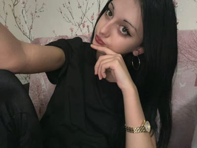A young woman with long, dark hair, dark eyebrows, winged eyeliner, and red lipstick is depicted in a medium shot, looking directly at the camera. She wears a black short-sleeved top and partially visible dark lower garments. Her right arm, extended and partially visible on the left, exhibits minor dermatological irregularities. Her left hand is raised, with fingers touching her chin and mouth. She wears a silver-colored hoop earring in her visible left ear and a gold-colored wristwatch with a metal band on her left wrist. The background consists of a wall covered in wallpaper featuring a repeating pattern of dark tree branches adorned with pink blossoms. A light pink, cushioned surface is partially visible behind and to her right. The scene is indoors.