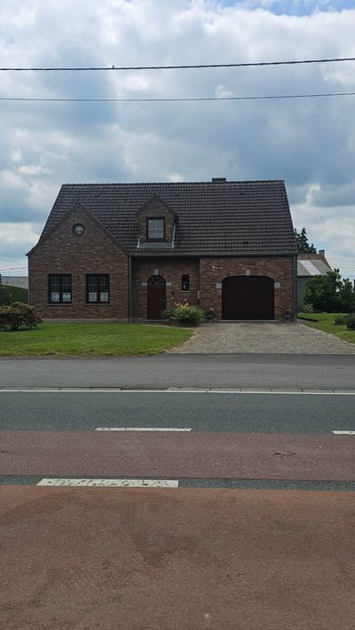 The image shows a brick house with a dark tiled roof, featuring a prominent arched garage door. There is a small lawn in front of the house, bordered by a paved driveway. A street with pedestrian crossing markings is visible in the foreground. The sky is overcast with clouds. The house is situated in Boussu-lez-Walcourt, Belgium, indicated by external data. No people or animals are visible in the scene.