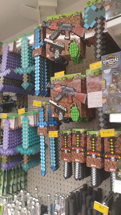 The image shows a retail display of Minecraft-themed toy weapons in a store. The items are primarily pixelated swords, axes, and guns, made of foam or plastic, packaged on cardboard backing designed to resemble the game's blocky aesthetic. Several price tags are visible, indicating prices in Euros, with one reading "E 4,99 л." and another "E 1,49 л." The display is arranged vertically on a pegboard wall. In the foreground, there are rows of other toys, including what appear to be silver-tipped staffs and dark, cylindrical objects, possibly foam batons or handles. The lighting suggests an indoor retail environment. The specific location is a store in Pylaia Municipal Unit, Greece.