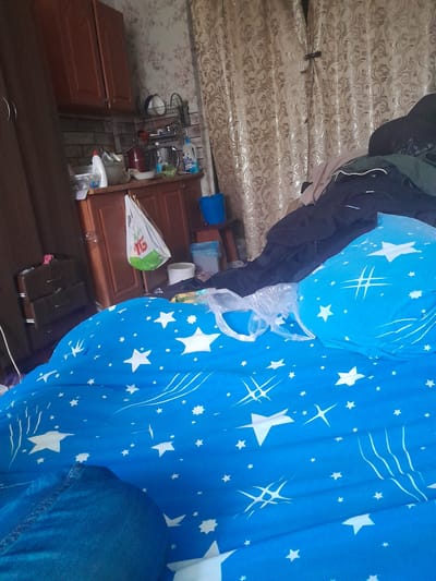 The image depicts an indoor scene, likely a domestic setting in Shostka, Ukraine. The primary subject is a large, bright blue fabric adorned with a pattern of white stars and streaks, taking up a significant portion of the right side of the frame. This fabric appears to be a duvet cover or similar bedding. To the left of the blue fabric, a dark, possibly black, garment is draped, hinting at clothing.
Behind these items, a collection of objects are visible, suggesting a cluttered or lived-in space. There are wooden cabinets with reddish-brown doors. On top of these cabinets, various household items are arranged, including a metal rack holding bowls, bottles, and what appears to be a sponge. There are also white plastic containers and a white plastic bag with a green logo that is partially visible. The background also features patterned wallpaper.
The lighting suggests it is daytime, but no specific time of day can be determined. There are no clear indicators of weather conditions. The overall impression is one of a casual, possibly storage or utility area within a home. No people are directly visible in the frame, but the presence of clothing and household items suggests human activity. The image is oriented with the top of the frame to the left.