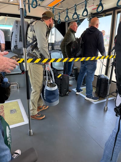 The image shows the interior of a shuttle or bus, likely at an airport. Several passengers are visible, along with their luggage. In the foreground, a person wearing a camouflage jacket and a green cap is seated. They have a large grey duffel bag beside them. To their left, another person in a green quilted jacket is standing. Further back, a person wearing a black hoodie and blue jeans is standing, with a black suitcase at their feet. The scene appears to be indoors, with a bright light suggesting daytime. The floor is dark grey, and there are silver poles for passengers to hold onto. Yellow and black striped hazard tape is visible along a pole. The overall atmosphere seems to be one of transit, with people waiting or preparing to board. There is no visible text in the image.