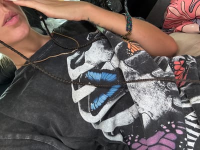 The media file shows a close-up, rotated view of a person wearing a black t-shirt with a large graphic print of butterflies. The t-shirt appears to be made of a distressed or washed fabric. The graphic includes purple, blue, and orange butterflies, some with black and white patterns on their wings. The person has braided hair that is draped across the t-shirt. A thin gold chain necklace is visible around the person's neck, and a colorful beaded bracelet is worn on their wrist. The person's arm and lips are also visible. The background is out of focus and appears to be an indoor setting with some fabric elements visible. The lighting suggests it is daytime, but there are no specific cues about the weather or a precise location beyond the mention of Bắc Yên, Vietnam, which is not evident from the image itself. The overall impression is casual and personal, focusing on the clothing and accessories. There is no visible text in the image.