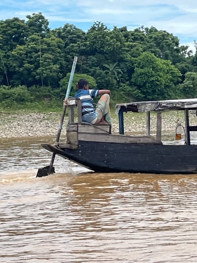 A person is rowing a boat on a river in Jaflong, Bangladesh. The person is wearing a blue and white striped shirt and patterned pants, and is seated on the boat. The boat is dark colored with a wooden structure. The river water is brown and appears murky. In the background, there is a lush green forested area on the riverbank, with some rocky terrain visible. The sky is partly cloudy.