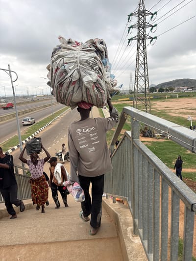 In Angwan Sayawa, Nigeria, a group of individuals are depicted on a pedestrian overpass. The central figure, a man, carries a large, tightly bound bundle of what appears to be used cement bags on his head. He is wearing a grey long-sleeved shirt with various logos including "Cigna," "AT&T," and "Citizen." Another man walks behind him carrying a smaller bundle on his head. To the left, a woman wearing a patterned skirt and a purple top carries a dark bag on her head. Two other individuals, a man and a woman, are also visible on the stairs. In the background, a highway with traffic, a power pylon, and a grassy field with goalposts are observed. The sky is overcast.