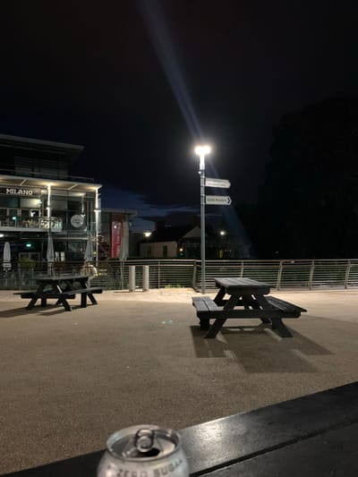 The image depicts an outdoor scene in Dublin 16, Ireland at night. A bright street lamp illuminates an area with restaurants, including one named "Milano" and signs pointing to "dlr Mill Theatre". There are metal benches and tables present, suggesting an outdoor dining area. An empty, crushed soda can lies discarded on the ground. People are vaguely visible in the illuminated restaurant. The overall environment suggests a commercial district at night.