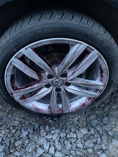 A close-up photograph taken in Zarvantsi, Ukraine, shows the front passenger side wheel of a vehicle parked on a gravel surface. The wheel features a dark rubber tire with a visible "GENERAL 4S" marking on its sidewall, mounted on a silver multi-spoke alloy rim. The center cap of the rim prominently displays the Volkswagen logo. A reddish-purple liquid, consistent with an iron-reactive wheel cleaning product, is observed splattered and dripping across the spokes and into the inner barrel of the rim. The ground beneath the wheel is composed of numerous irregular gray gravel pieces interspersed with sparse dry vegetation. No individuals or other active subjects are visible in the frame.