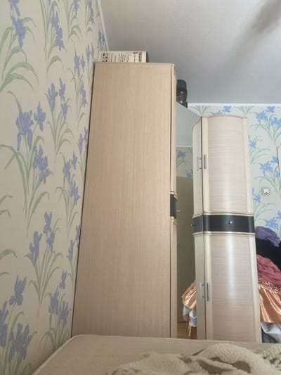 The image shows an interior scene. A tall, light-colored wood wardrobe with a vertical wood grain texture is positioned against a wall with floral wallpaper featuring blue irises and green leaves. To the right of this wardrobe, there is another, narrower wardrobe with rounded corners and a similar light wood finish, accented with a dark band near the middle and metal handles. This wardrobe appears to be a mirrored or glass-fronted unit, as a reflection of the room behind the viewer can be seen. Clothing, including pink and orange garments, is visible spilling out of the right side of the scene, suggesting the wardrobes may be partially open or used for storage of laundry. The ceiling is visible at the top, and a portion of a bed or mattress covered with a white textured throw is in the foreground. The scene is likely within a residential dwelling in Kovrov, Russia.