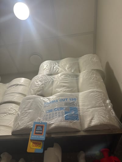 The image depicts an indoor storage or retail area, showcasing multiple bundles of white paper products stacked on a dark-colored shelf. Each bundle consists of several individually plastic-wrapped rolls, further enclosed in clear plastic. The frontmost bundle features a label reading "FARMA CUT 130" and "Papirnate brisalce Handtowel Handtücher," along with a barcode and QR code. Attached to this bundle are two hanging price tags: an upper blue tag displaying the "HOFER" logo and the Slovenian text "Več kot fer. HOFER.", and a lower yellow tag marked "HOFER CENE". These labels indicate the products are consistent with items sold at a Hofer supermarket within Slovenia. The specific location within Radovljica, Slovenia, cannot be forensically confirmed from the image itself. The overhead environment consists of a white suspended acoustic tile ceiling with grid lines and two circular flush-mounted light fixtures, one of which is brightly illuminated. A plain, light-colored wall is visible on the right side of the frame. A lower shelf, partially visible beneath the main display, holds indistinct white objects and a red plastic item, possibly a container. No individuals are present in the scene.