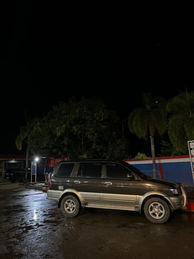 At night in Seunebobaru, Indonesia, a dark gray SUV is parked on a wet, reflective asphalt surface. The SUV is positioned in the middle of the frame, facing towards the right. Its headlights are off. The ground is illuminated by a single overhead light source that creates a bright reflection on the wet pavement. Behind the SUV, a red and blue fence runs horizontally, separating the parking area from a row of buildings. Lush green trees, including palm trees with distinctive fronds, are visible behind the fence, and a dark, dense tree with broad leaves is located to the left of the SUV. The sky is completely black, indicating it is nighttime. In the background, on the far left, another dark-colored vehicle is partially visible.