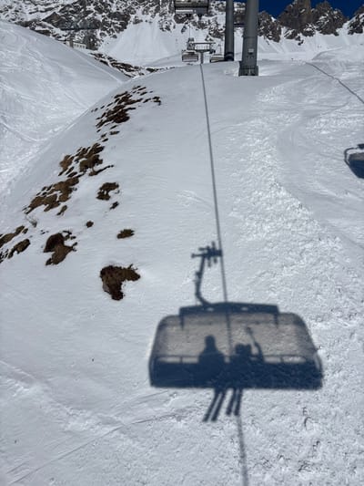 This image depicts a high-angle view from a ski lift, overlooking a snow-covered mountain slope within a ski resort in Klösterle, Austria.
In the foreground, the distinct dark shadow of a ski lift cabin or chair is projected onto the snow-covered ground below. Within this shadow, two primary human silhouettes are visible, seated, with vertical lines extending downwards, consistent with skis. A third, smaller and less distinct silhouette appears adjacent to one of the main figures. The shadow of the lift's connecting cable and the apparatus holding the cabin is also present.
The midground features a snow-covered slope interspersed with irregular patches of exposed, brown-colored ground or vegetation. Multiple ski tracks are etched into the snow, particularly noticeable on the right side of the frame.
The background shows the upward progression of the ski lift system, with several gray metal poles supporting cables from which empty lift chairs are suspended. In the far distance, rugged mountain peaks, partially covered in snow with exposed rocky faces, rise against a clear, bright blue sky. A small building, likely a lift station or mountain facility, is visible on the left slope in the upper background.
The scene is illuminated by direct sunlight, creating sharp, prominent shadows, indicative of a clear day.