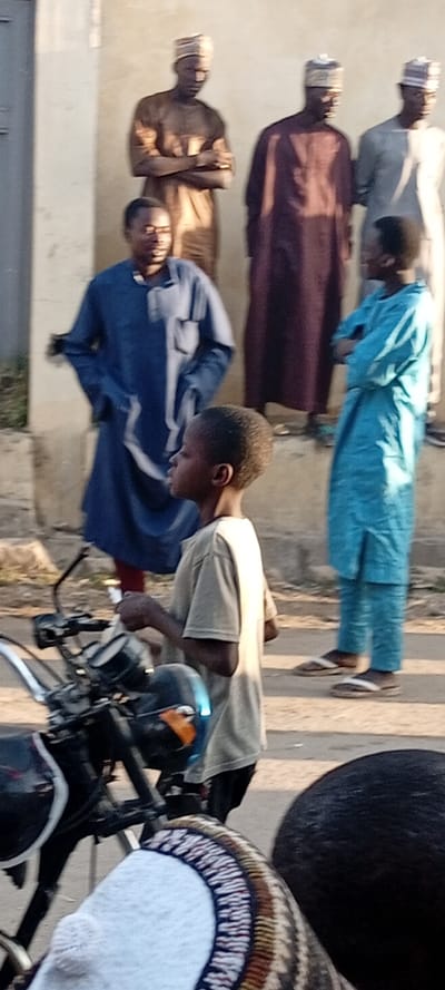 The image depicts an outdoor street scene in Kabuga, Nigeria, under natural daylight. In the immediate foreground, the handlebars and a portion of the seat of a black motorcycle are visible. A young male, approximately 8-12 years old, with short dark hair, wearing a light tan short-sleeved t-shirt and dark trousers, stands to the right of the motorcycle, facing left in profile. He holds a small, light-colored object in his right hand.

In the midground, from left to right:
1.  A male in a dark blue long-sleeved kaftan, standing with hands in his pockets, looking towards the right.
2.  A male in a light blue long-sleeved kaftan and matching trousers, wearing sandals, standing with arms crossed, looking towards the left.

In the background, against a plain, light-colored wall, three adult males are partially visible, all wearing traditional caps. From left to right:
1.  A male in a light brown kaftan, standing with arms crossed.
2.  A male in a dark maroon kaftan.
3.  A male in a light grey kaftan.

The ground appears to be an unpaved, uneven surface. Shadows indicate a light source from the right side of the frame. The individuals are standing in various states of observation or interaction with each other and their surroundings.