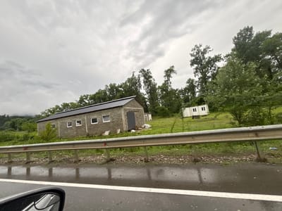 This media file depicts a cloudy, possibly rainy day scene along a road in the Nigogiti community of Georgia. The foreground shows a wet asphalt road with white lane markings and reflections from the overcast sky. A silver guardrail runs horizontally across the frame, separating the road from the area beyond.
Behind the guardrail, a long, rectangular building constructed from cinder blocks is visible. It has a dark, sloped roof and several square windows with white frames. A large, dark double door is situated on the side of the building. To the right of this building, a smaller, white prefabricated structure with two square windows is also present.
The background consists of a grassy slope with scattered trees and dense foliage. The terrain rises to form hills in the distance, which are partially obscured by clouds or mist, suggesting a mountainous or hilly region. The overall atmosphere is somber, likely due to the overcast weather. No people are visible in the media file, and no text is present. The image appears to be taken from a moving vehicle, as indicated by the car's side mirror in the lower left corner of the frame.