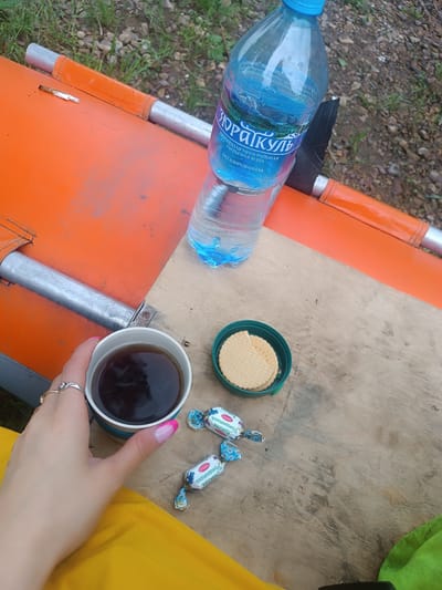 The image depicts a close-up, overhead view of a personal break outdoors. A hand with manicured nails, adorned with two rings, holds a white mug filled with dark liquid, likely coffee or tea. The mug rests on a light-colored wooden surface, which is adjacent to an orange object, possibly part of a boat or recreational equipment. To the right of the mug are two individually wrapped candies, their wrappers featuring blue floral patterns and Russian text. Further to the right, a green plastic lid contains two round, light-colored biscuits. Behind the wooden surface, a clear plastic bottle of water with a blue label is visible, partially obscured by the orange object. The background consists of gravel and green grass. The scene appears to be set in a natural outdoor environment, potentially a park or riverside area. The provided metadata indicates the location as Idrisovo, Russia.