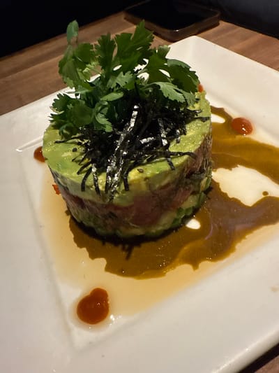 A multi-layered food stack, presented cylindrically, occupies the center of a white rectangular plate. The stack consists of alternating layers: an uppermost layer of diced green avocado, a middle layer of diced red protein, and a lower layer of diced green avocado. The top is garnished with fresh green cilantro and shredded black nori seaweed; small black sesame seeds are visible on the avocado surface. A dark amber-brown sauce is distributed around the base of the stack on the plate. Two distinct, small, circular drops of an orange-red condiment are present on the plate, separate from the main sauce, positioned to the upper right and lower left of the food stack. The white plate rests on a wooden table. The background is dark and out of focus, with a dark rectangular object visible on the table surface in the upper right. The scene is located in Sunrise, United States of America.
