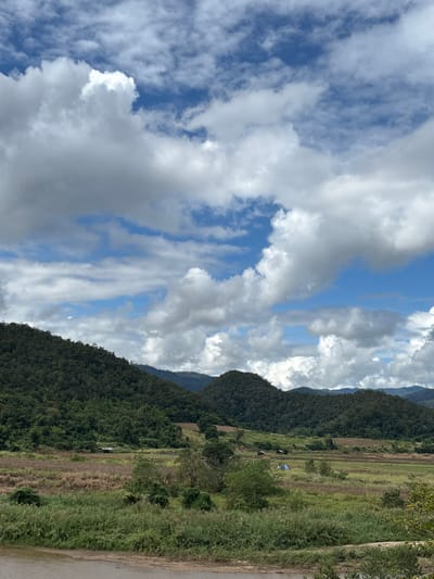 A landscape scene depicts a sky filled with dynamic white and gray clouds, contrasting with patches of blue sky. A verdant, mountainous region occupies the right side of the frame, densely covered with trees. At the base of the mountains, there's a mix of grassy fields and cultivated areas, possibly agricultural land. A narrow strip of road is visible at the bottom right corner.