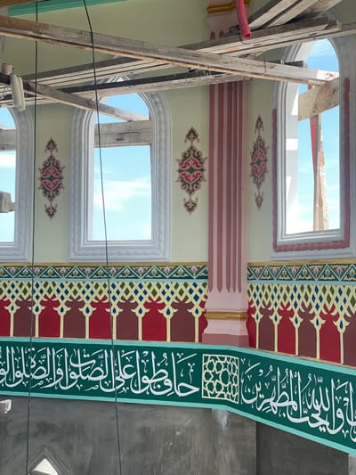 This image captures the interior of a mosque under construction or renovation in Kumbang, Indonesia. The scene is dominated by a brightly lit wall featuring intricate Islamic architectural and decorative elements.

The upper section of the wall is painted a pale yellow, topped by a lighter mint green band near the ceiling. Multiple arched windows, currently without glass, punctuate this section, revealing a clear blue sky with sparse white clouds. The windows are framed by ornate white moldings with fluted details. Between these windows, and adjacent to a light pink fluted pillar with vertical darker pink stripes and a gold base, are symmetrical decorative motifs in red, pink, and dark green, featuring curvilinear patterns. Overhead, wooden scaffolding planks and beams are visible, indicating ongoing construction. Several thin black wires hang vertically in the foreground, likely for electrical or other installations.

Below the windows, a horizontal frieze begins with a multi-colored geometric lattice pattern in yellow, dark green, blue, and red. Beneath this, a band of repeated red and dark brown arch-like shapes is painted. The lowest visible decorated section is a prominent green band with white Arabic calligraphy, interspersed with a geometric star pattern. Below this finished section, the wall transitions to an unfinished, raw gray concrete surface, further highlighting the ongoing work. No individuals are present in the scene.