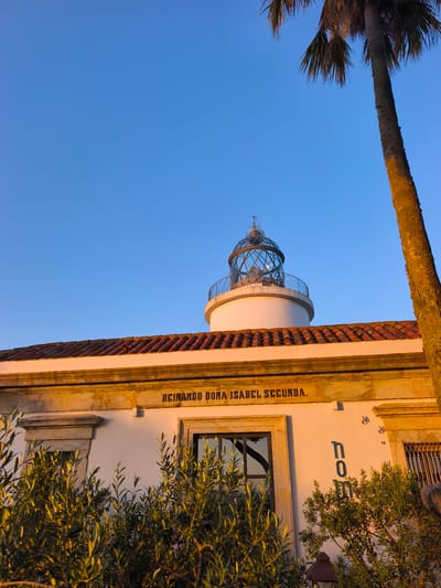 The image shows the lighthouse of Sant Sebastià, located on the coast of Palafrugell, Spain. The lighthouse is white with a red tiled roof and a glass lantern at the top. A palm tree stands to the right of the lighthouse. The sky is clear blue, and the sun is setting, casting a warm glow on the scene. The text "REINANDO DOÑA ISABEL SECUNDA" is inscribed on the building. There are also green bushes in the foreground. The overall scene is peaceful and scenic.