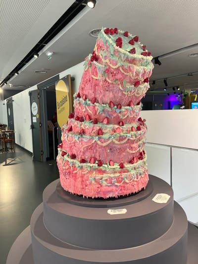 A large, multi-tiered sculpture resembling a melting cake, predominantly pink with dripping white and light blue frosting, adorned with numerous red fruit representations and small blue bows, is displayed on a tiered, dark grey cylindrical pedestal. A white label on the pedestal states "NE PAS TOUCHER / DON'T TOUCH." The sculpture is situated within an indoor exhibition space characterized by a dark floor, white walls, and a white perforated ceiling fitted with black track lighting. In the background, a doorway features a prominent yellow elliptical sign reading "En cuisine / In the Kitchen." Further down the hallway, another door is labeled "Entrée / Entrance." To the far left, a few chairs and tables are partially visible. To the right of the cake, a different exhibit area with distinctive purple and blue lighting is visible beyond a white partition. This scene is located in the Cité de la Gastronomie et du Vin in Dijon, France. No people are visible.