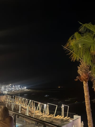 The image captures a night scene in Saint Paul's Bay, Malta. In the upper left corner, the leaves of a palm tree are visible. Along the right side of the image, there's a row of closed beach umbrellas with white frames, indicating a beachfront establishment, possibly a restaurant. Lights are strung out along the beach, suggesting a populated area, likely for evening dining or relaxation. A person can be seen in the foreground. The background is a dark sky, indicating nighttime. The view is from an elevated position, perhaps from a balcony or terrace.