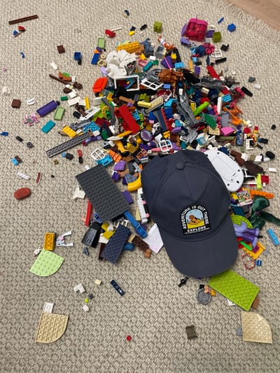 A dark blue baseball cap with a circular patch is placed on a carpeted surface, surrounded by a large assortment of scattered Lego bricks and other small toys. The patch on the cap reads "ADVENTURE IS OUT THERE EXPLORE LIFE IS BETTER IN THE MOUNTAINS". The Lego pieces are of various colors, shapes, and sizes, including larger base plates and smaller individual bricks. Some Lego figures and accessories are also visible. The carpet is a neutral, textured beige. The scene appears to be a typical disarray of children's toys. The image does not contain information that would indicate its location in the city of Cahul, Moldova, or any specific geographical information.