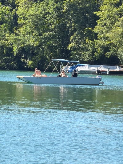 This image captures a serene scene on the Rhine River in Eglisau, Switzerland. In the foreground, the clear, blue water shimmers with ripples. In the middle ground, a long, narrow boat with a light-colored hull is positioned horizontally. The boat has a blue canopy supported by metal poles.
On the bow of the boat, a shirtless person is lying down, facing away from the camera. To their right, another person is seated and appears to be interacting with someone else in the boat. Further back in the boat, another individual is visible, wearing a dark top. The boat has a license plate that reads "ZH-7756."
In the background, a dense line of lush green trees forms a natural backdrop. Behind the boat and further in the distance, there are other docked boats, some covered with white tarpaulins, suggesting a marina or waterfront area. The overall impression is one of a tranquil summer day on the river, with people enjoying leisure time on the water.