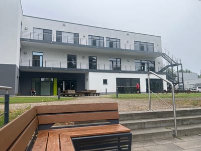 The image shows the exterior of a modern, multi-story building in Teltow, Germany. The building has a white facade with dark trim and numerous windows, many with horizontal blinds. Balconies with metal railings are visible on the upper floors. An external metal staircase leads to the top floor on the right side of the building. In the foreground, a wooden bench with a dark metal frame sits on a paved area with concrete steps leading up to it. Behind the bench, a gravel path is present, with a patch of grass and some trees further in the background.
On the ground floor of the building, there are glass doors and large windows, some of which appear to be entrances. In front of these entrances, there are wooden picnic tables. To the left, a person is seated on a white chair outside the building, looking at a book or tablet. To the right, a person wearing a red shirt and dark shorts stands on the gravel path in front of the building. Several cars are parked in the background. The sky is overcast, suggesting a cloudy day. The overall scene depicts an institutional or commercial building with outdoor seating and recreational elements.