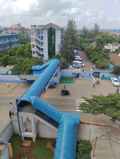 An elevated, slightly oblique view depicts an urban scene in the Hospital ward, Kenya, under an overcast sky. The foreground is dominated by a covered pedestrian bridge with a blue corrugated metal roof and blue railings, descending diagonally from the lower left across a two-lane asphalt road, then extending horizontally left. The road features a white pedestrian crosswalk.
To the left, a multi-story building with a blue and white exterior is partially visible, surrounded by trees. In the mid-background, a taller, modern multi-story building stands with a light grey facade, green-tinted glass sections, external yellow staircases, and a green roof. Further right in the background are residential structures with light-colored walls and brown roofs.
Approximately 10-12 individuals are present. Three persons walk towards the left entrance of the pedestrian bridge. One person in white attire stands near a barrier further left. On the right side of the road, near an entrance, approximately 5-7 individuals are walking or standing, with one person in dark uniform attire positioned near a white parked sedan. Other vehicles include a black three-wheeled auto-rickshaw on the road, several white vans/minibuses parked in a lot behind the bridge section, and motorcycles near the left bridge base. Numerous trees and green shrubs are dispersed throughout the environment, particularly along the road and between buildings.