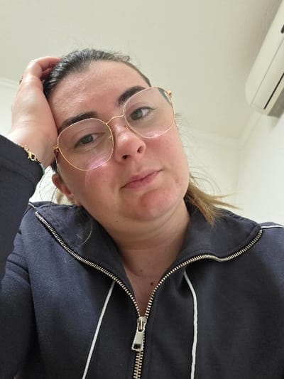 A close-up, indoor photograph taken in Negorci, North Macedonia, features a female individual positioned centrally. The subject has fair skin, dark hair pulled back, and wears large, round, gold-rimmed eyeglasses. She is dressed in a dark blue zip-up jacket with a silver zipper closure and white piping visible on the lapel. Her right hand is raised, with the palm resting against her forehead and fingers on the side of her head. Her gaze is directed forward towards the camera, maintaining a neutral expression. The background consists of a plain white wall, with a white, wall-mounted air conditioning unit visible in the upper right portion of the frame.