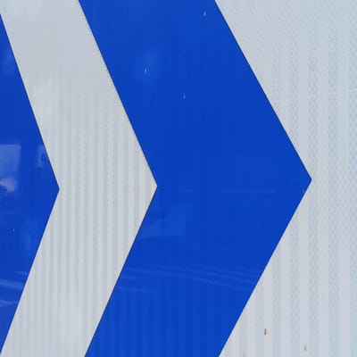 The image presents a close-up, abstract view of a reflective surface, likely part of a traffic sign or barrier. The primary subjects are alternating blue and white chevron-like patterns. The blue sections are solid and reflective, forming angular shapes that point towards the right side of the frame. The white sections are also highly reflective and display a fine, repeating grid or honeycomb texture, characteristic of retroreflective material.
No specific activity or event is depicted, as the image is a static shot of an object. Small imperfections, such as tiny white specks on the blue areas and brownish spots on the white sections, are visible, suggesting some wear or environmental exposure. There is no visible text. The time of day, weather, and specific location cues (beyond the provided Chancheng District, China context) are not discernible from the image itself. The composition highlights the geometric design and the material's property of reflecting light for visibility.