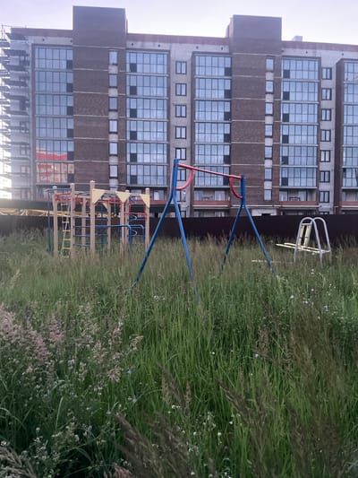 The image depicts a residential building and a playground in Laishevo, Russia. The building is a multi-story structure with a facade of brick and large, grid-like windows. On the right side of the image, a playground is visible, featuring a swing set with red and blue frame elements and a climbing structure with yellow, blue, and red components. The playground is surrounded by tall, green grass and wildflowers, suggesting an outdoor setting during daytime. The sky appears overcast, contributing to a diffused lighting. The overall scene is calm and ordinary, typical of a residential area. No people are visible in the image.