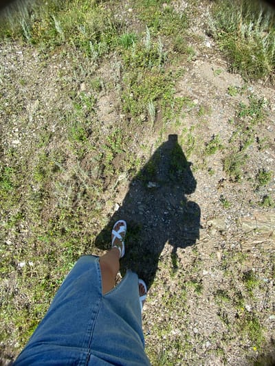 This is a first-person, downward-facing, wide-angle shot of a person standing on gravelly ground covered with sparse grass and small plants. The location is Yuldybayevo, Russia. The person is wearing a denim skirt and white sandals. A shadow of the person, cast by a bright sun, is visible on the ground. The shadow suggests the person is holding a camera or phone at arm's length. The overall environment appears to be an outdoor, possibly rural or semi-rural setting, with dry, rocky soil and green vegetation.