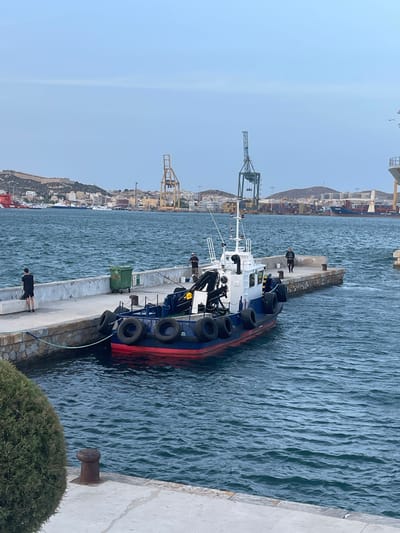 In Cartagena, Spain, a small, blue and red boat with a white superstructure is docked at a concrete pier. Several black fenders are attached to the boat's side. Two individuals stand on the pier near the boat. One, wearing a black t-shirt and shorts, is facing away from the camera and towards the water, appearing to be fishing. Another person, in a dark shirt and pants, stands further down the pier, also facing away. A green trash receptacle is present on the pier. In the background, the city of Cartagena is visible with buildings and hills, and several large cranes dominate the skyline. The water is choppy, and the sky is overcast.