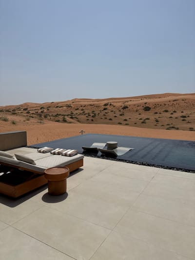 An infinity pool is situated at the edge of a luxurious desert resort in Ras Al-Khaimah, United Arab Emirates. The pool's dark blue water reflects the clear, pale blue sky. Adjacent to the pool, a minimalist patio is laid with large, light-colored rectangular tiles. A wooden chaise lounge with beige upholstery and striped towels is placed on the patio, accompanied by a small, round wooden side table. In the background, vast sand dunes stretch towards the horizon, dotted with sparse, dry vegetation, indicative of a desert landscape. The scene conveys a sense of serene luxury and isolation in a desert environment.