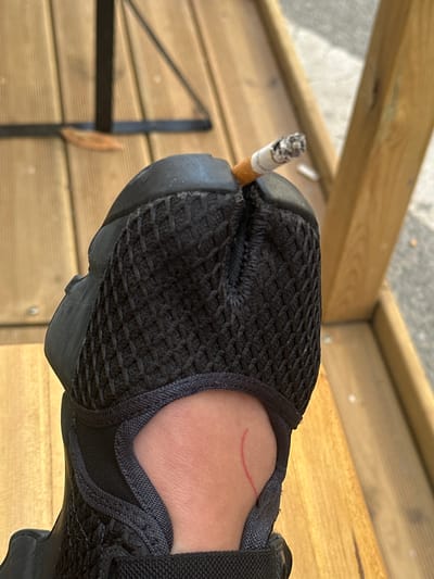 A close-up photograph shows a black athletic shoe with a lit cigarette wedged into the toe. The shoe is being worn, evidenced by the exposed ankle and foot visible in the frame. A red mark is visible on the wearer's ankle. The background features wooden planks, potentially a deck or outdoor seating area. A black metal object, possibly part of outdoor furniture, is also partially visible. The image does not provide sufficient information to determine the location within Saint-Ouen-sur-Seine, France.